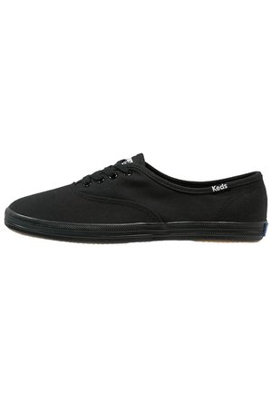 Baskets Keds en toile noire avec lacets à l'avant, semelle en caoutchouc profil bas et petite étiquette de marque près du talon sur fond blanc.