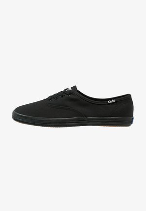 Zapatilla Keds de lona negra con cordones delanteros, suela de goma baja y pequeña etiqueta de marca cerca del talón sobre fondo blanco.