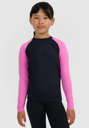 Girl standing with black and pink long-sleeve shirt and black pants against a plain light gray background.