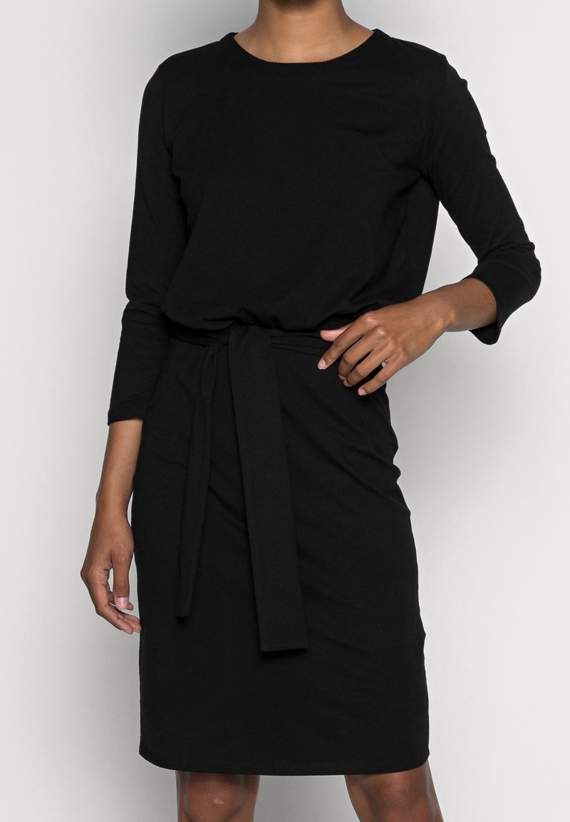 Femme portant une robe noire longueur genou avec des manches trois-quarts et une ceinture en tissu nouée à la taille, debout devant un fond uni.