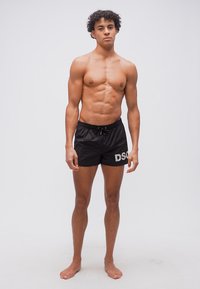 Jeune homme aux cheveux bouclés, debout pieds nus, portant un short de bain noir DSQ, sur un fond clair uni.