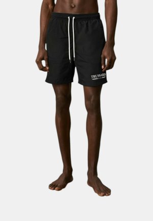 Persona scalza che indossa pantaloncini da bagno neri con laccetti bianchi e logo "Trussardi" sulla gamba sinistra su uno sfondo semplice.