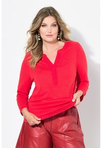 Rotes Langarmshirt mit V-Ausschnitt und gerippten Details, kombiniert mit hochtaillierten, glänzenden roten Lederhosen. Glatte Textur insgesamt.