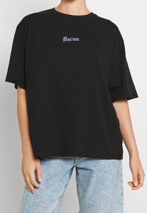 Persona vistiendo una camiseta negra holgada con el texto púrpura "Karma" en el pecho y jeans de mezclilla azul claro, de pie contra un fondo liso.