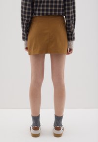 Gonna mini in suede marrone, vita aderente, texture liscia. Abbinata a una camicia a quadri e calzini grigi, con sneakers bianche e marroni.