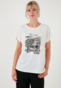 Hvid kortærmet t-shirt med et sort og sølv grafisk design, der indeholder abstrakte mønstre og tekst. Blødt stof, afslappet pasform, rund halsudskæring.