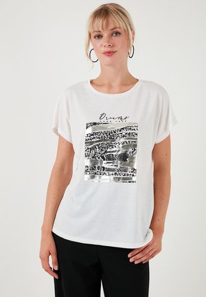 T-shirt blanc à manches courtes avec un design graphique noir et argent présentant des motifs abstraits et du texte. Tissu doux, coupe décontractée, col rond.