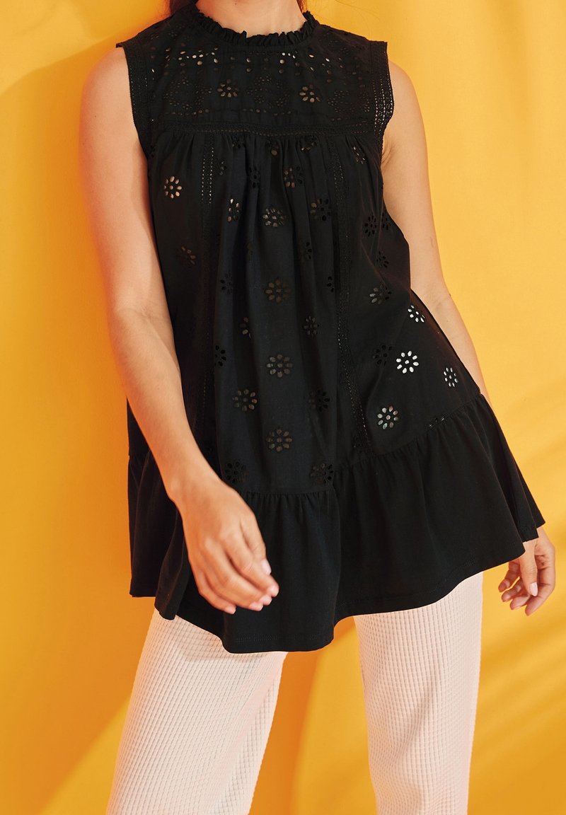 Personne portant un haut noir sans manches en œillets avec des découpes florales et un pantalon côtelé crème sur un fond jaune.