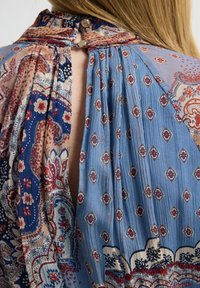 Espalda de mujer que muestra una blusa de alto cuello de múltiples estampados con diseños florales y geométricos en azul, rojo y beige, abrochada con dos botones.