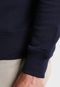 Il felpa blu marina presenta un polsino aderente, un orlo a costine e una texture liscia. Il tessuto sembra leggero e elastico.