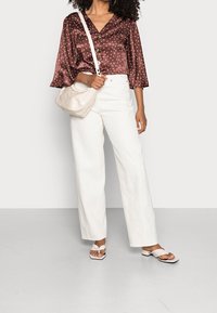 Brun blus med prickar, V-ringning och trekvartsärmar, i kombination med vita vida byxor och beige sandaler. Håller en krämfärgad väska.