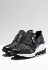 Sneakers neri con pannelli in rete, una cinghia con zip sulla parte anteriore e una suola intermedia bianca. Superficie testurizzata e suola in gomma.