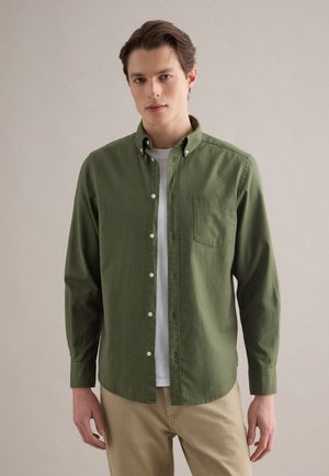 Junger Mann, der ein olivgrünes Hemd über einem weißen T-Shirt und beige Hosen trägt, vor einem schlichten hellen Hintergrund stehend.