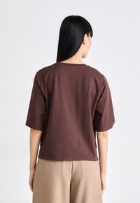 Donna con lunghi capelli neri che indossa una maglietta marrone a maniche corte e pantaloni beige, in piedi di spalle su uno sfondo neutro.