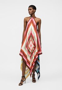 Γυναίκα που φορά πολλαχρωμη τουαλέτα με λουράκι στον λαιμό, με ρίγες σε σχήμα διαμαντιού και ασύμμετρο τελείωμα, συνδυασμένη με μαύρα πέδιλα με ψηλό τακούνι.