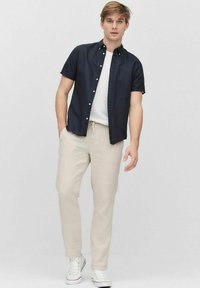 Kortärmad marinblå skjorta över en vit t-shirt, kombinerad med ljusbeiga byxor och vita sneakers, mot en neutral bakgrund.