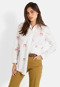 Chemise blanche à boutonnage avant avec un nœud rouge et des motifs en script, assortie à un pantalon beige et une ceinture à imprimé léopard. Tissu lisse et design classique.