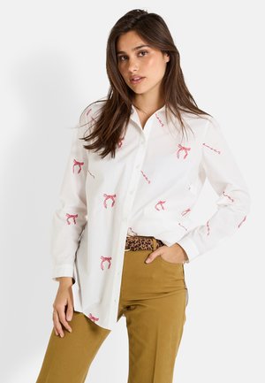 Chemise blanche à boutonnage avant avec un nœud rouge et des motifs en script, assortie à un pantalon beige et une ceinture à imprimé léopard. Tissu lisse et design classique.