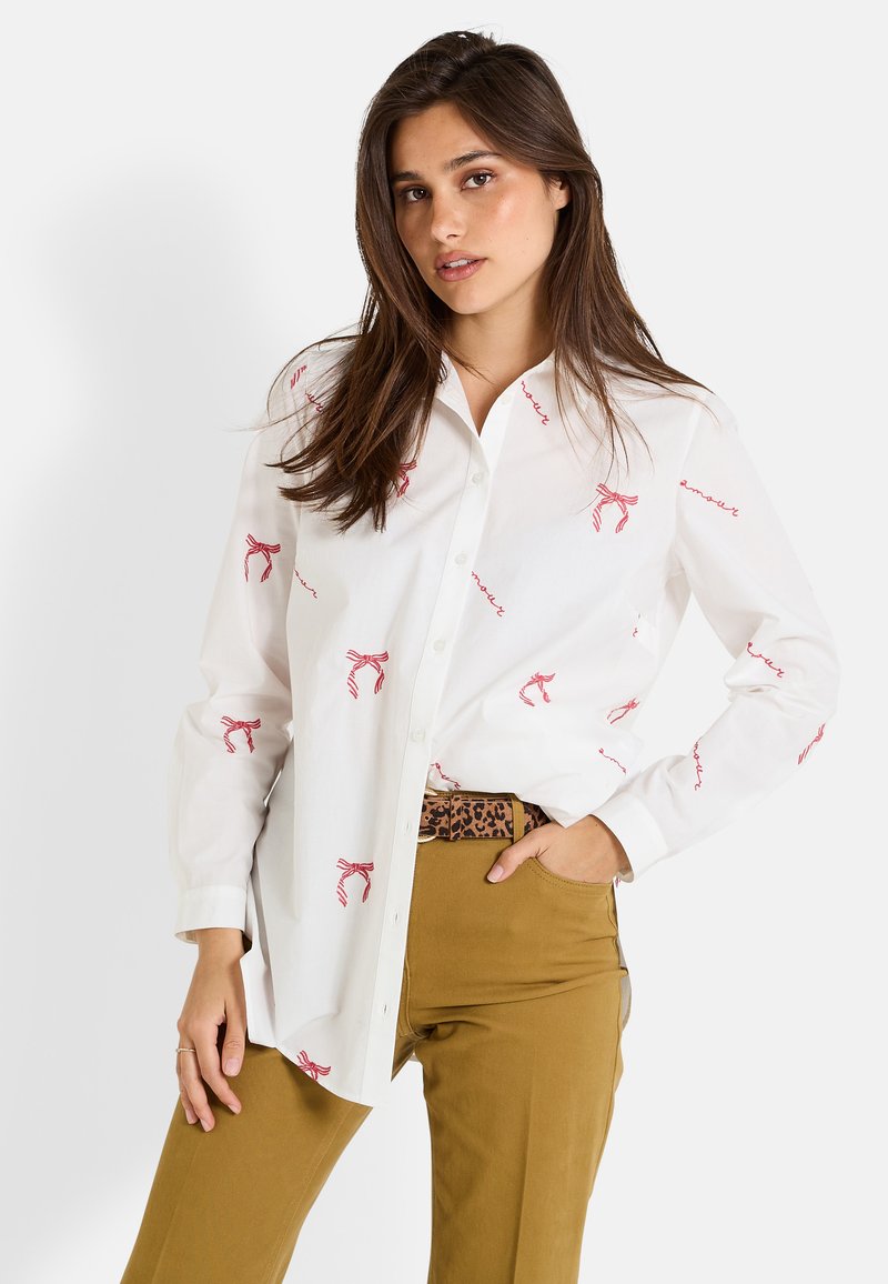 Chemise blanche à boutonnage avant avec un nœud rouge et des motifs en script, assortie à un pantalon beige et une ceinture à imprimé léopard. Tissu lisse et design classique.