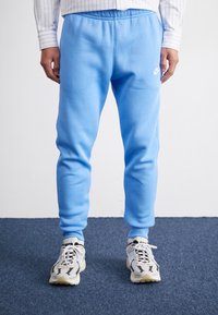 En person som bär ljusblå träningsoverallsbyxor och vita sneakers står på en mörkblå matta mot en enfärgad bakgrund. Händerna avslappnat vid sidorna.