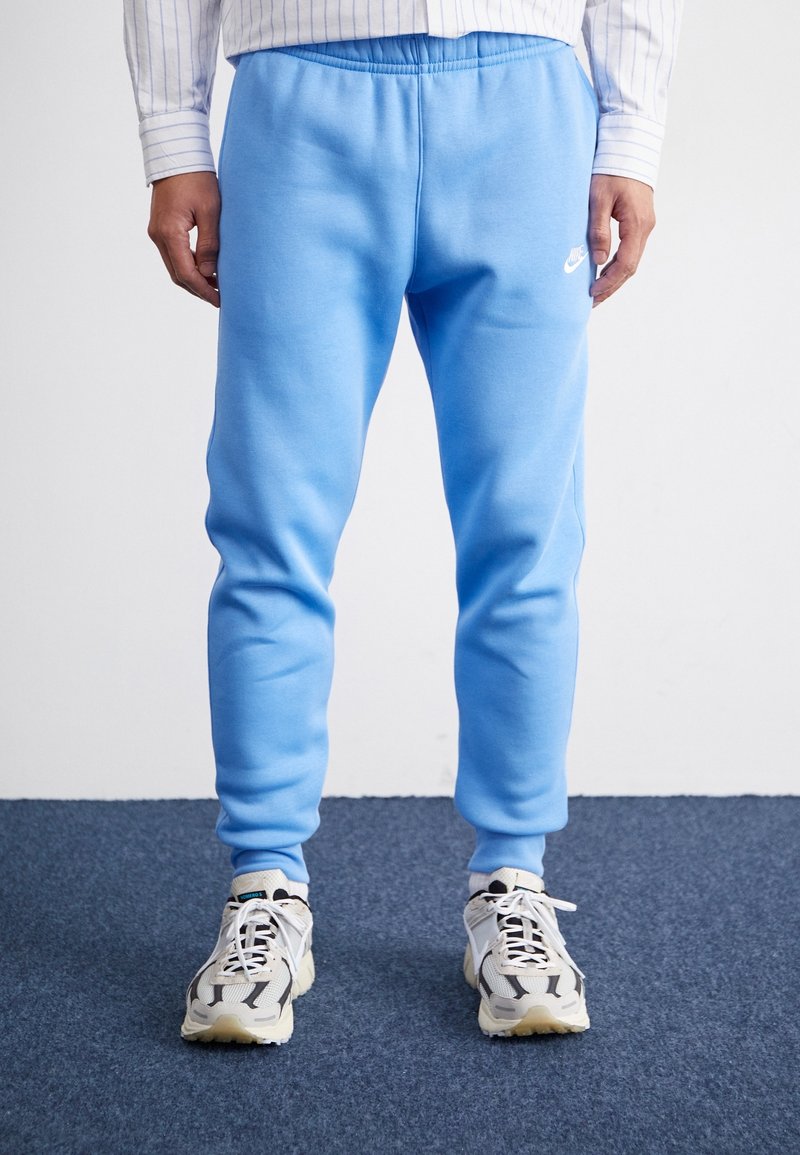 En person som bär ljusblå träningsoverallsbyxor och vita sneakers står på en mörkblå matta mot en enfärgad bakgrund. Händerna avslappnat vid sidorna.