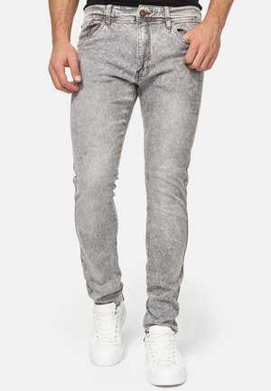 Homme portant un jean slim gris acid-wash et des baskets blanches, debout, une main dans la poche sur un fond uni.
