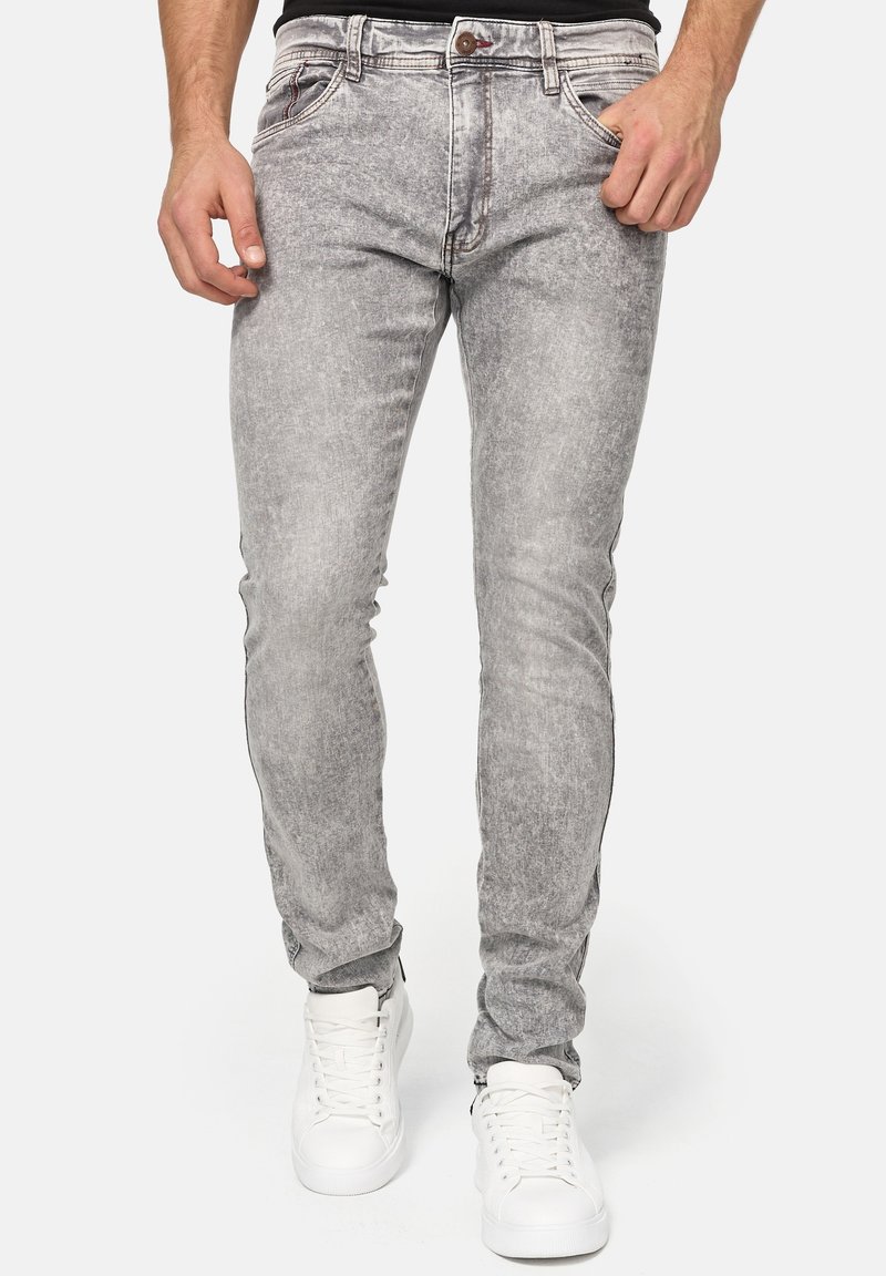 Homme portant un jean slim gris acid-wash et des baskets blanches, debout, une main dans la poche sur un fond uni.