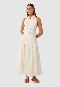 Vestido longo branco com gola alta, fecho na frente e cintura com cinto. Apresenta pregas verticais e saia fluida. Sandálias em tom claro. 