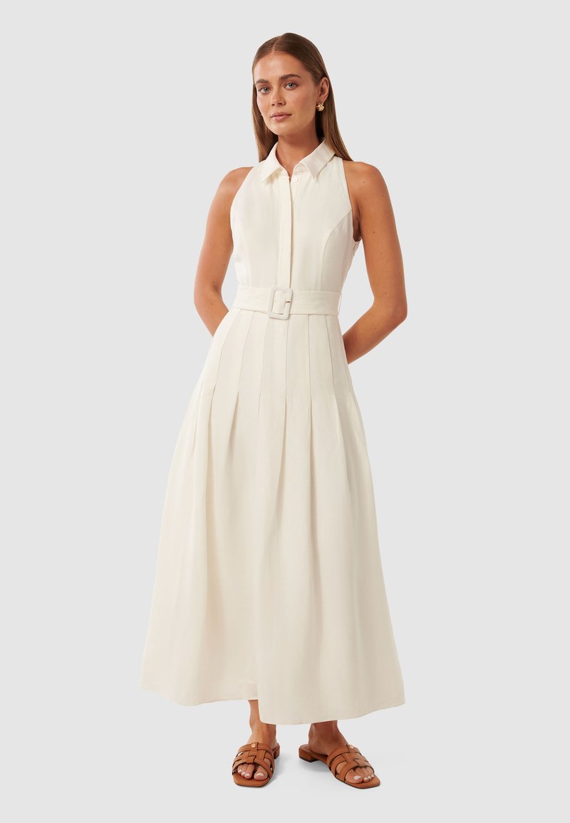 Vestido longo branco com gola alta, fecho na frente e cintura com cinto. Apresenta pregas verticais e saia fluida. Sandálias em tom claro. 