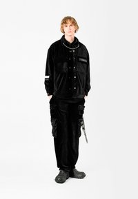 Chaqueta de pana negra con botones a presión, grandes bolsillos delanteros y detalles en plata, combinada con pantalones a juego y calzado negro texturizado.