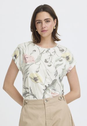 Õhuke pruunide õlgadeni juustega naine, kes kannab kuldseid rõngaskõrvarõngaid, valget lillemustriga pluusi ja beežika vöökohaga pükse, seisab käed selja taga.