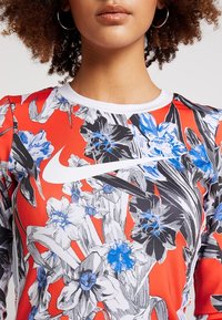 Donna che indossa un top floreale rosso con grandi fiori blu e grigi e il logo bianco dello swoosh Nike sul petto.