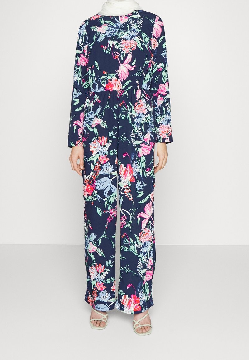 Kombinezon w kwiatowy wzór z głębokim granatowym tłem oraz żywymi różowymi, niebieskimi i zielonymi kwiatami. Długie rękawy i szerokie nogawki.