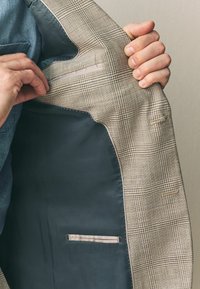 Interior de una chaqueta de traje de cuadros grises con un forro oscuro. Incluye un bolsillo interior con cremallera y un lazo de tela. Textura suave.
