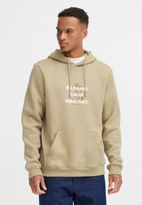 Sudadera color beige con un bolsillo frontal, que presenta un estampado gráfico que dice "EXPANDE TU MENTALIDAD" en colores contrastantes y cordones.