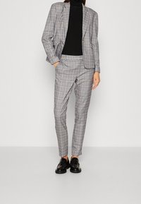 Szary garnitur w kratę z dopasowaną marynarką o strukturalnym kroju i obcisłymi spodniami. W połączeniu z czarnym golfem i czarnymi mokasynami. Z subtelnymi guzikami jako akcentami.