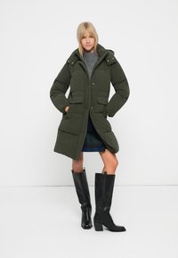 Olivenfarvet polstret jakke med hætte, to forlommer og knaplukninger, båret over en grå sweater og en blåternet nederdel, parret med sorte støvler.