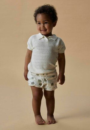 Polo de punto blanco con mangas cortas, combinado con pantalones cortos claros que presentan un sutil estampado animal, de pie descalzo contra un fondo neutro.