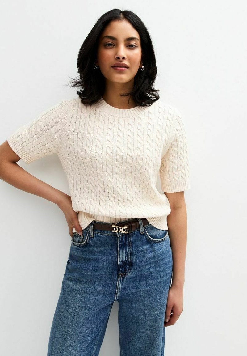 Kremowy sweter robiony na drutach z krótkimi rękawami, z teksturowanym wzorem w warkocz. Połączony z jeansami w kolorze niebieskim o wysokim stanie i brązowym paskiem.