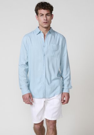 Light blue button-up shirt made of soft fabric with a chest pocket and long sleeves, paired with white shorts. Minimal design details.
