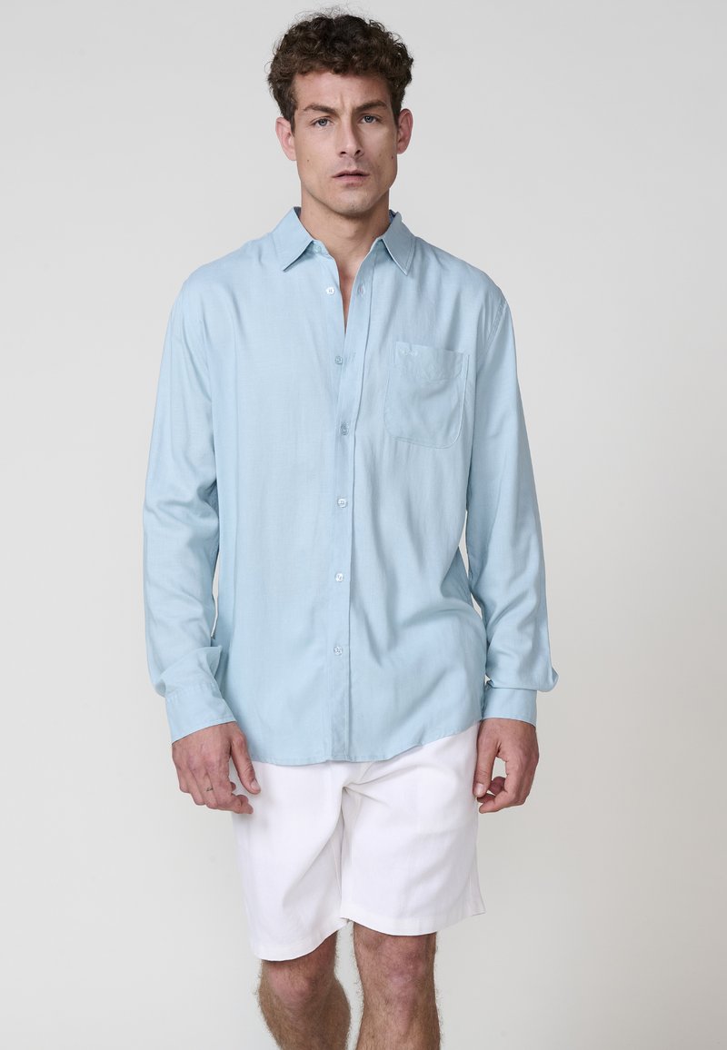Camicia azzurro chiaro con bottoni, realizzata in tessuto morbido, con taschino sul petto e maniche lunghe, abbinata a pantaloni corti bianchi. Dettagli di design minimalisti.