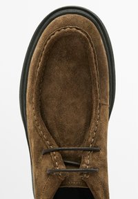 Chaussure en suède marron avec un bout arrondi, semelle en caoutchouc noir et deux lacets en cuir marron. Couture détaillée le long des bords.