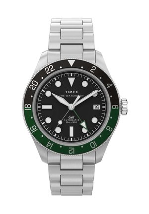 Nerūdijančio plieno Timex Waterbury GMT laikrodis su juodu ciferblatu, žalia ir juoda bezelė, šviečiančiais žymekliais, datos langeliu ir sidabriniu metaliniu apyrankiu.