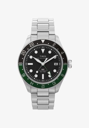 Hodinky Timex Waterbury GMT z nerezové oceli s černým ciferníkem, zeleno-černým rámečkem, luminiscenčními značkami, okénkem data a stříbrným kovovým náramkem.