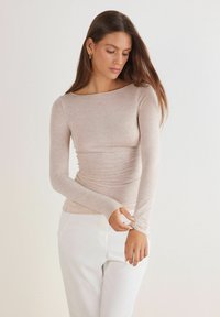 Långärmad ljusbeige topp med båthals och rynkad textur vid midjan, parat med vita byxor. Slät tyg.