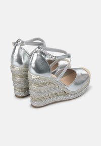 Sandali wedge metallici argento con cinturini incrociati e dettagli in juta intrecciata. Il wedge presenta intricate decorazioni argentate e una punta arrotondata.
