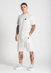 Weißes, gestreiftes T-Shirt mit kurzen Ärmeln und passender Shorts mit Kordelzug. Verfügt über ein kleines Logo-Patch. Blaue Turnschuhe mit weißen Punkten.
