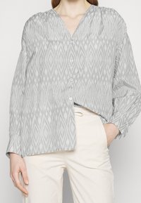 Blusa grigia a fantasia con scollatura a V, caratterizzata da un design geometrico, chiusura con bottoni e maniche lunghe. Indossata con pantaloni color crema.