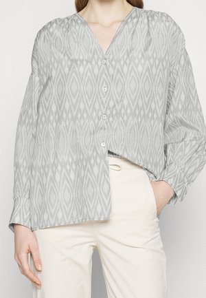 Blusa grigia a fantasia con scollatura a V, caratterizzata da un design geometrico, chiusura con bottoni e maniche lunghe. Indossata con pantaloni color crema.