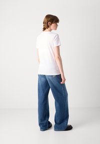 T-shirt bianco in cotone con maniche corte, abbinato a jeans blu di denim larghi. Lo sfondo è bianco uniforme, evidenziando l'outfit.
